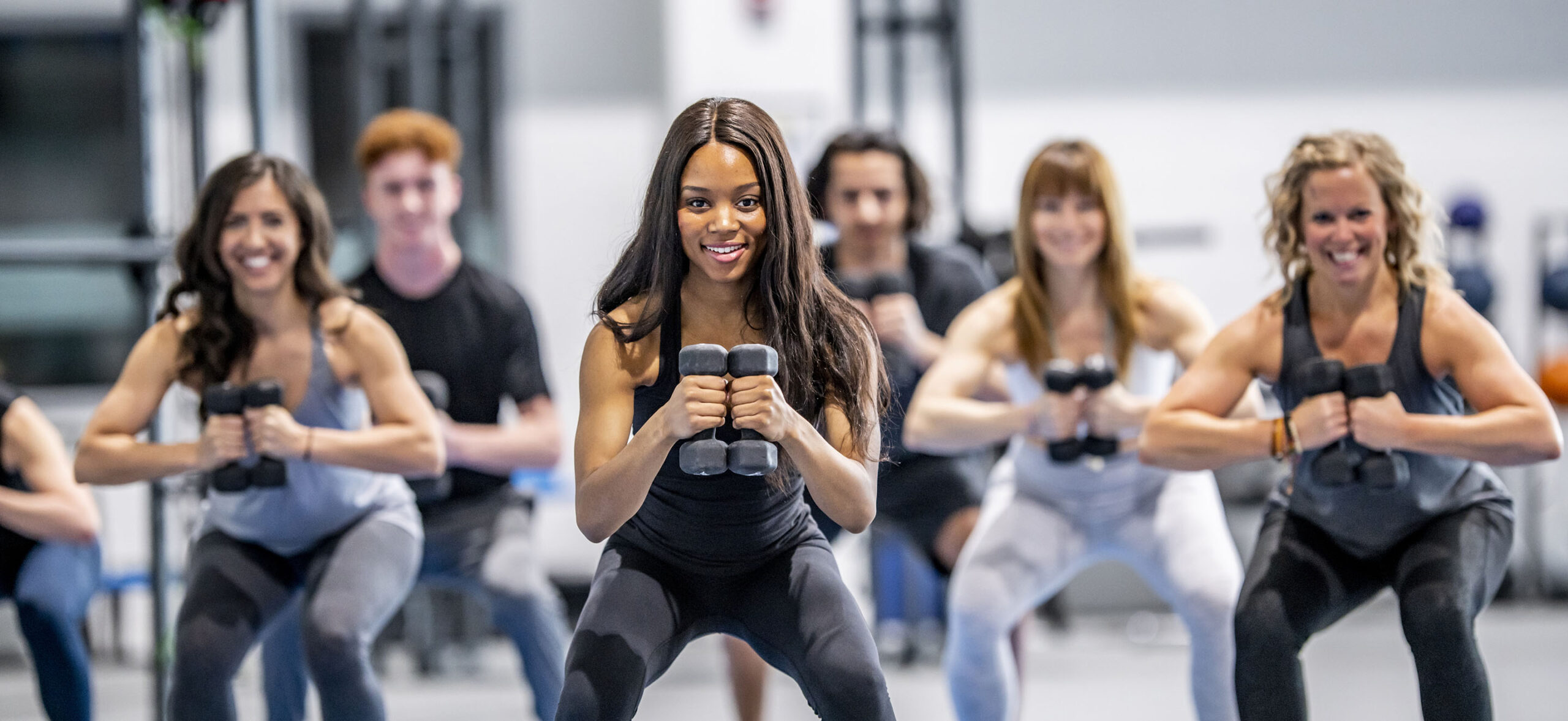 A multi-ethnic group of men and women are indoors in a gym. They are smiling while squatting and lifting dumbbells.