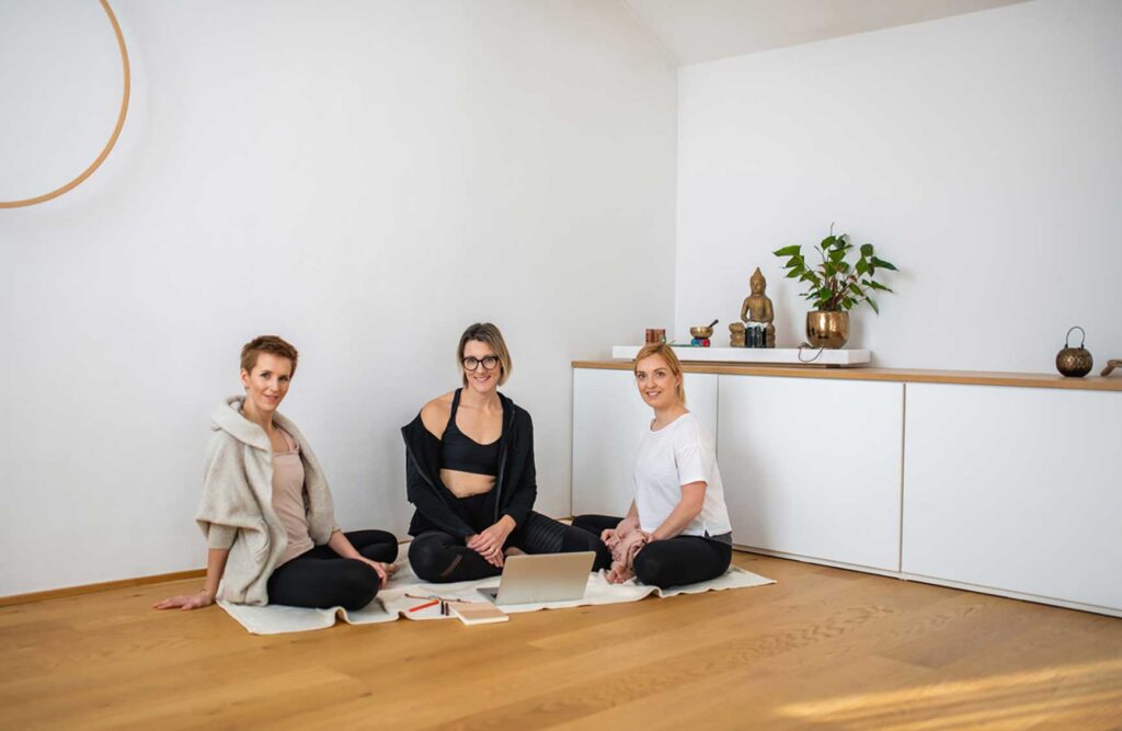 Full length front view of Caucasian women in early 30s sitting on floor in yoga studio and pausing from using laptop to smile at camera.