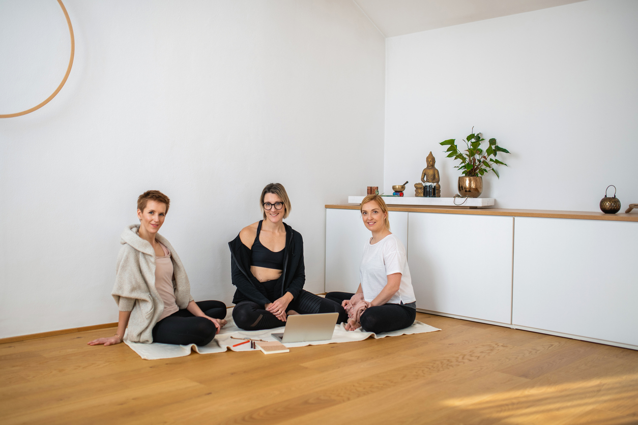 Full length front view of Caucasian women in early 30s sitting on floor in yoga studio and pausing from using laptop to smile at camera.