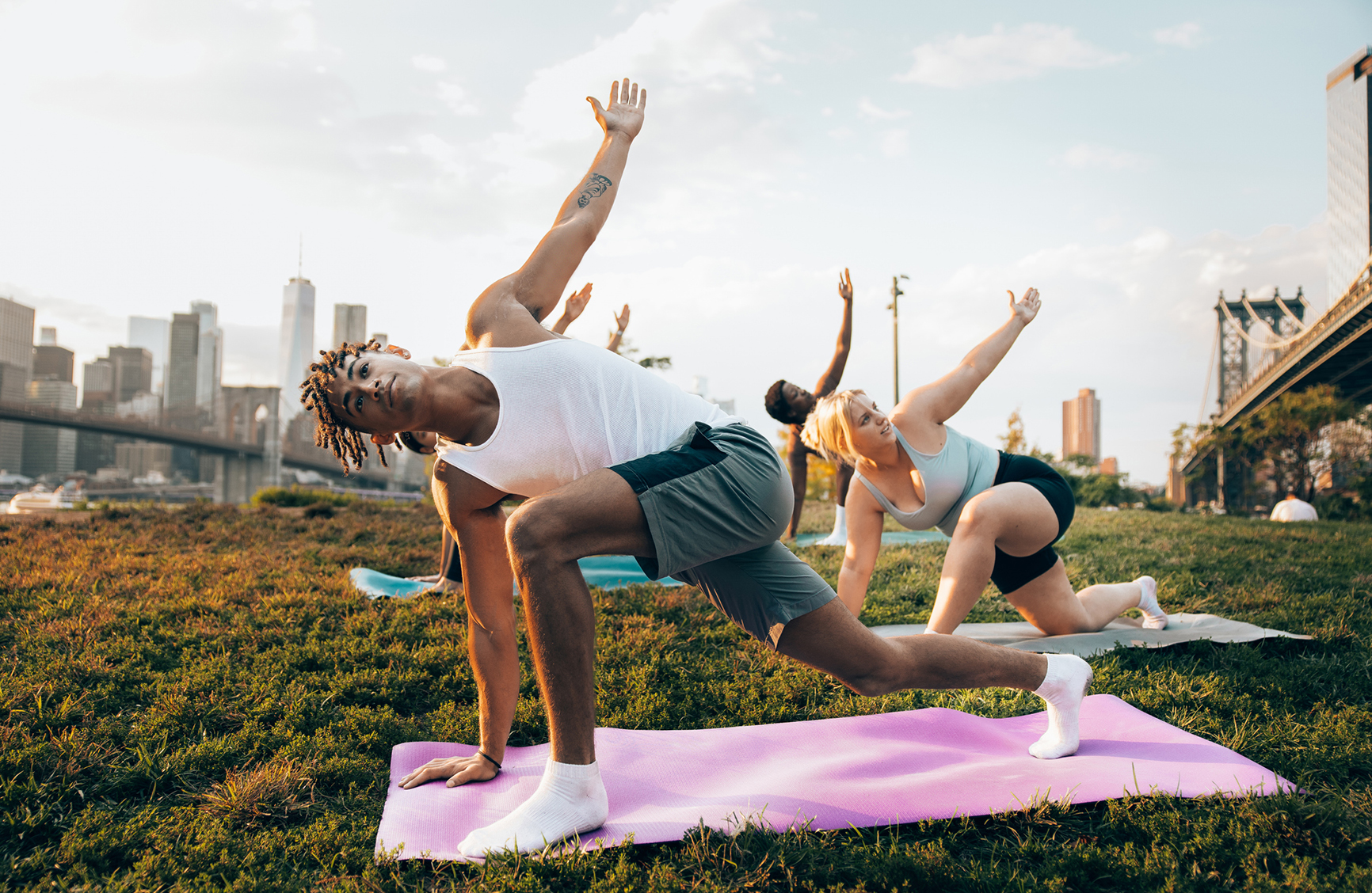 Group of young people attending to a yoga class outdoors at sunset with New York cityscape on their background. They are meditating and relaxing.