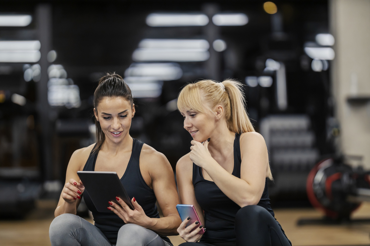 A happy female trainer is analyzing progress on tablet with sportswoman wile sitting in a gym.