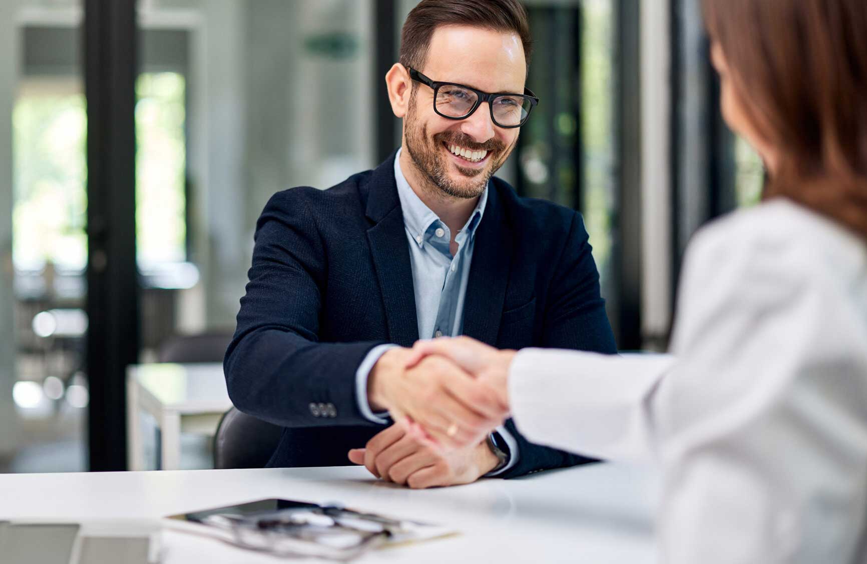 A smiling businessman greets a female doctor with a stethoscope on the office table and handshakes with her.