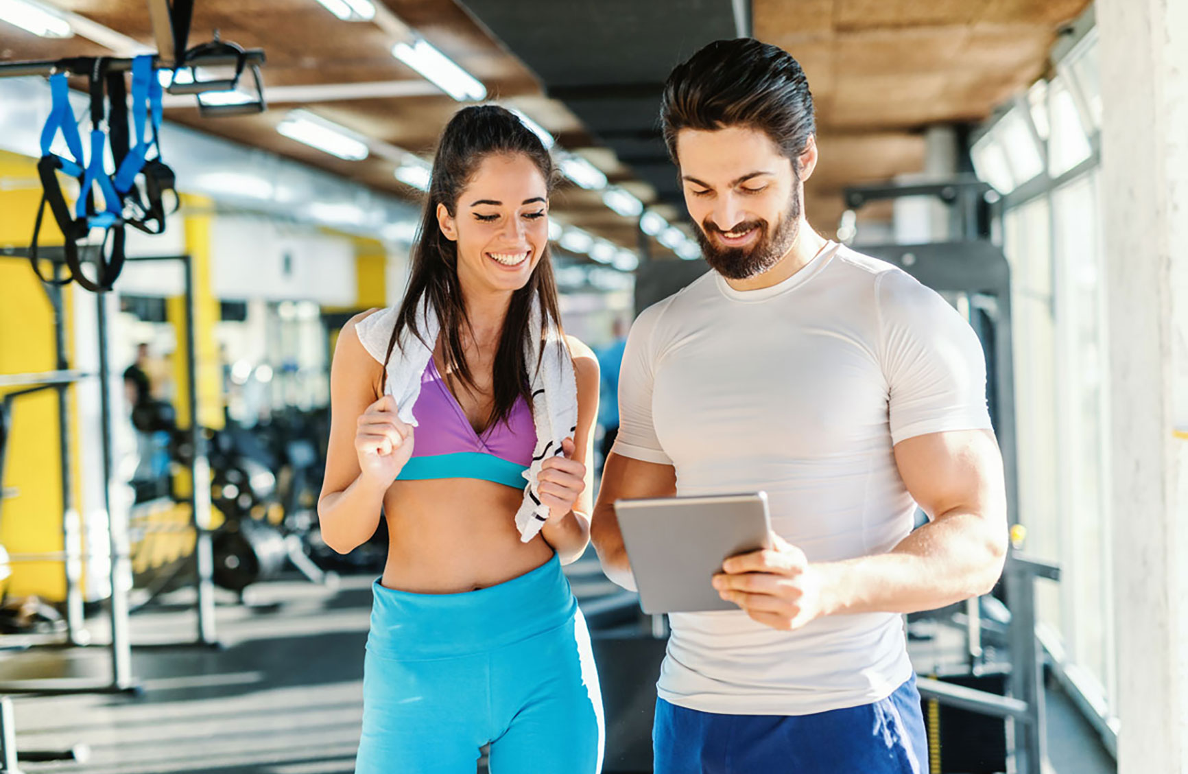 Bearded smiling personal trainer showing woman results of training on tablet. Woman standing next to him and looking at tablet. Gym interior.