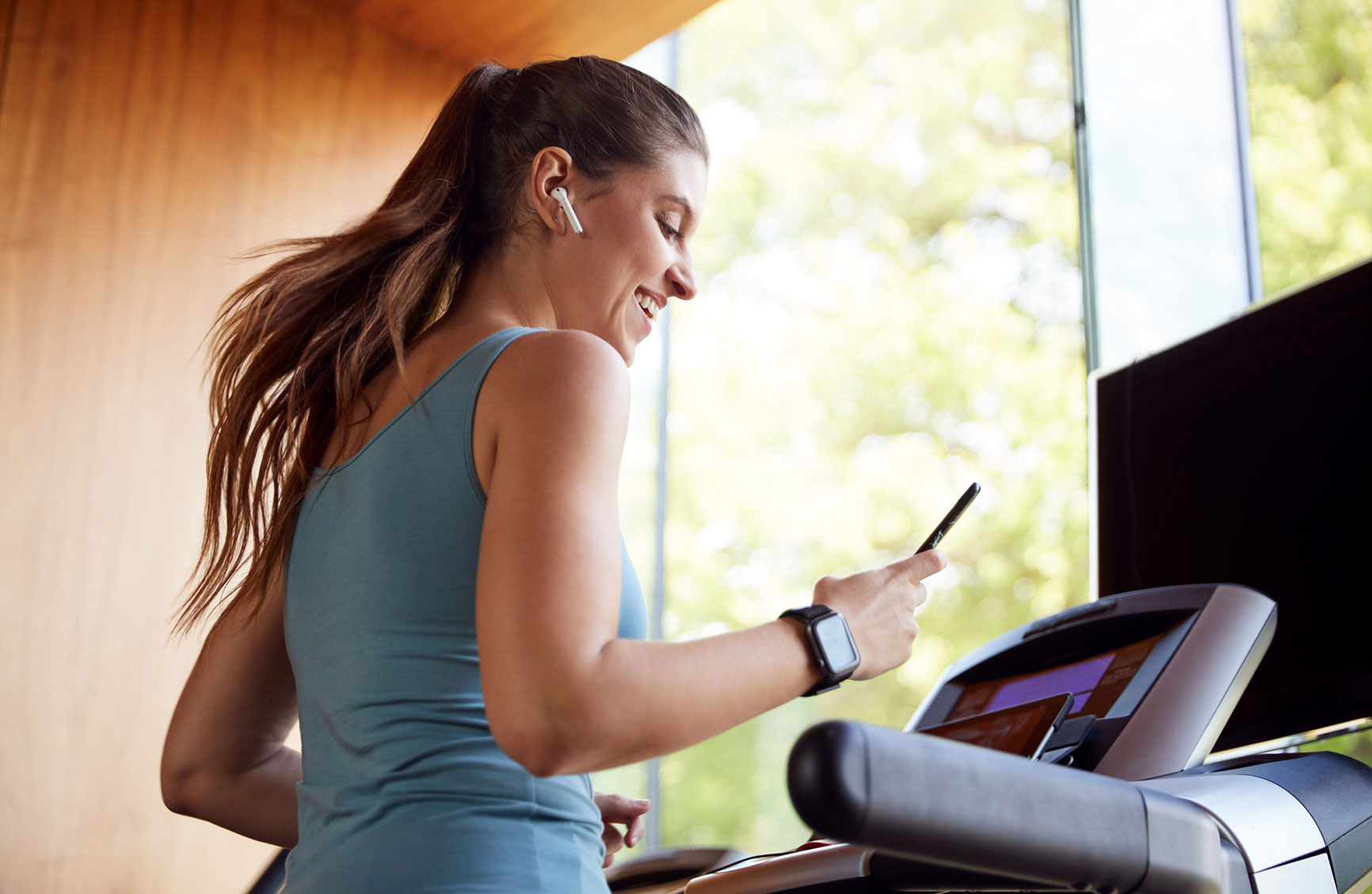 Woman Exercising On Treadmill Wearing Wireless Earphones And Smart Watch Checking Mobile Phone