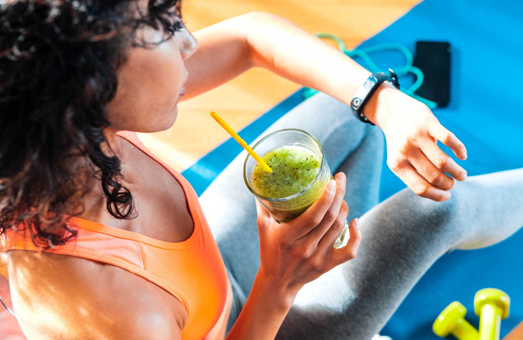 Sporty woman in sportswear training at home drinking fresh smoothie - Fit female athlete using smart watch to monitor her performance - Sport, food and technology concept.