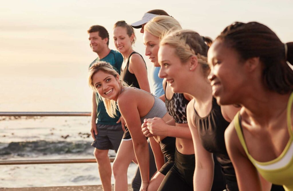 Group of young athletes in start position, focus on woman. Fit young people preparing for race along sea.