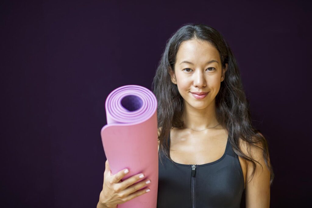 A lady smiling at the camera with a yoga mat.